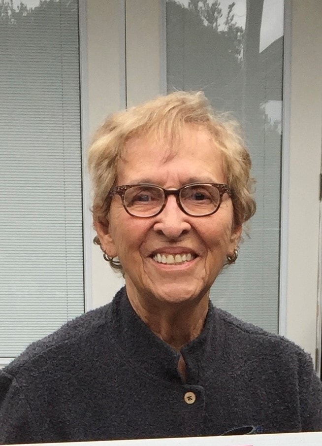 An older woman with short, light hair and glasses smiles at the camera, standing in front of a window with blinds.