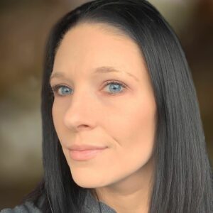 A woman with straight black hair and blue eyes looks at the camera with a neutral expression against a blurred background.
