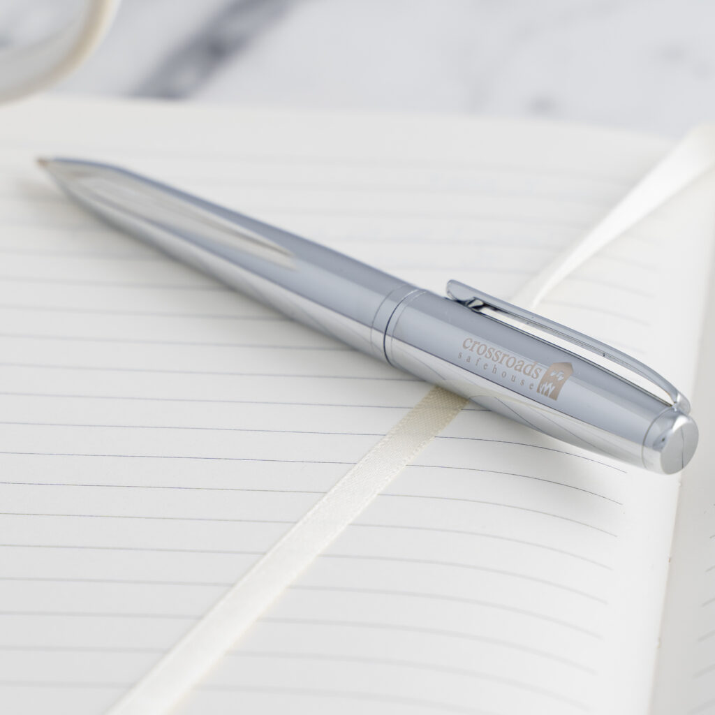 A silver pen rests on an open lined notebook with a white ribbon bookmark.