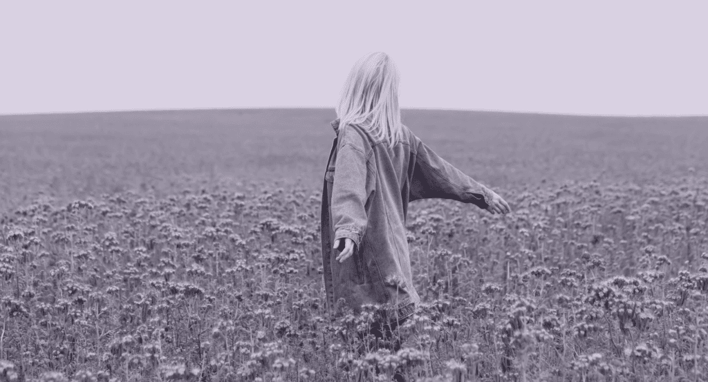 A person with long hair, wearing an oversized jacket, stands in a field of wildflowers with arms outstretched, facing away from the camera.