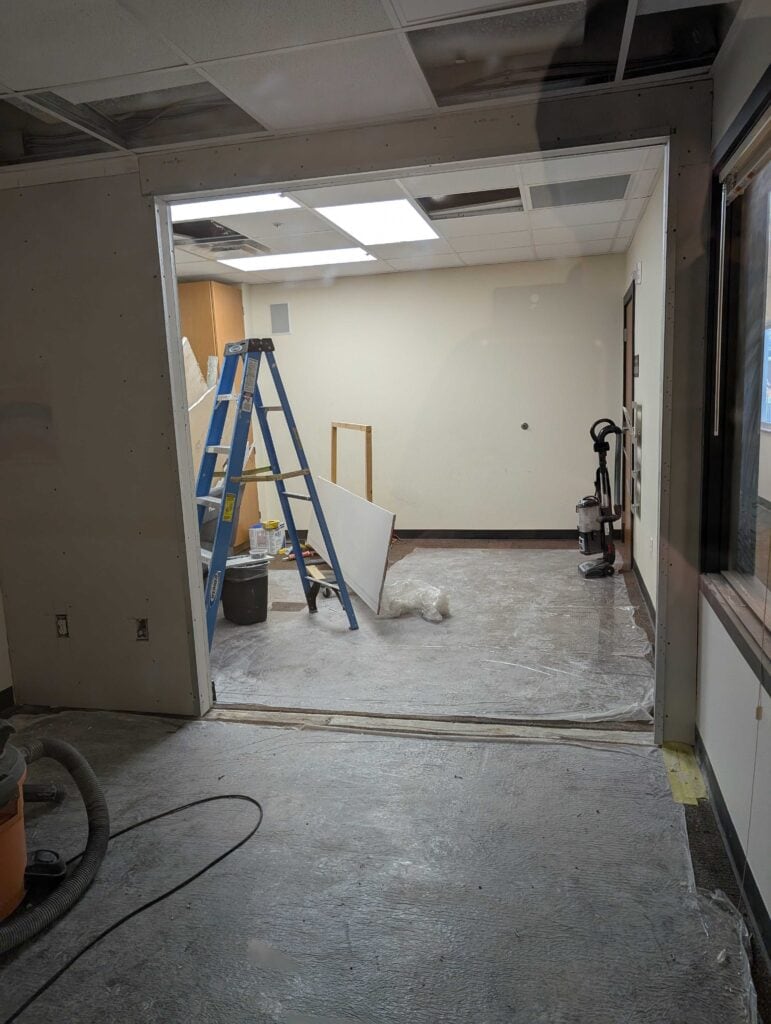 A room under renovation with a blue ladder, construction materials, vacuum cleaner, and unfinished walls and ceiling.