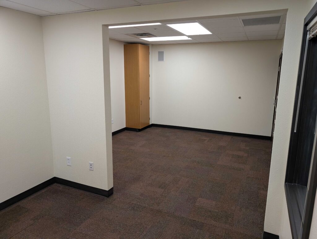 Empty office room with beige walls, brown carpet, fluorescent ceiling lights, a wooden cabinet in the corner, and visible electrical outlets on the left wall.