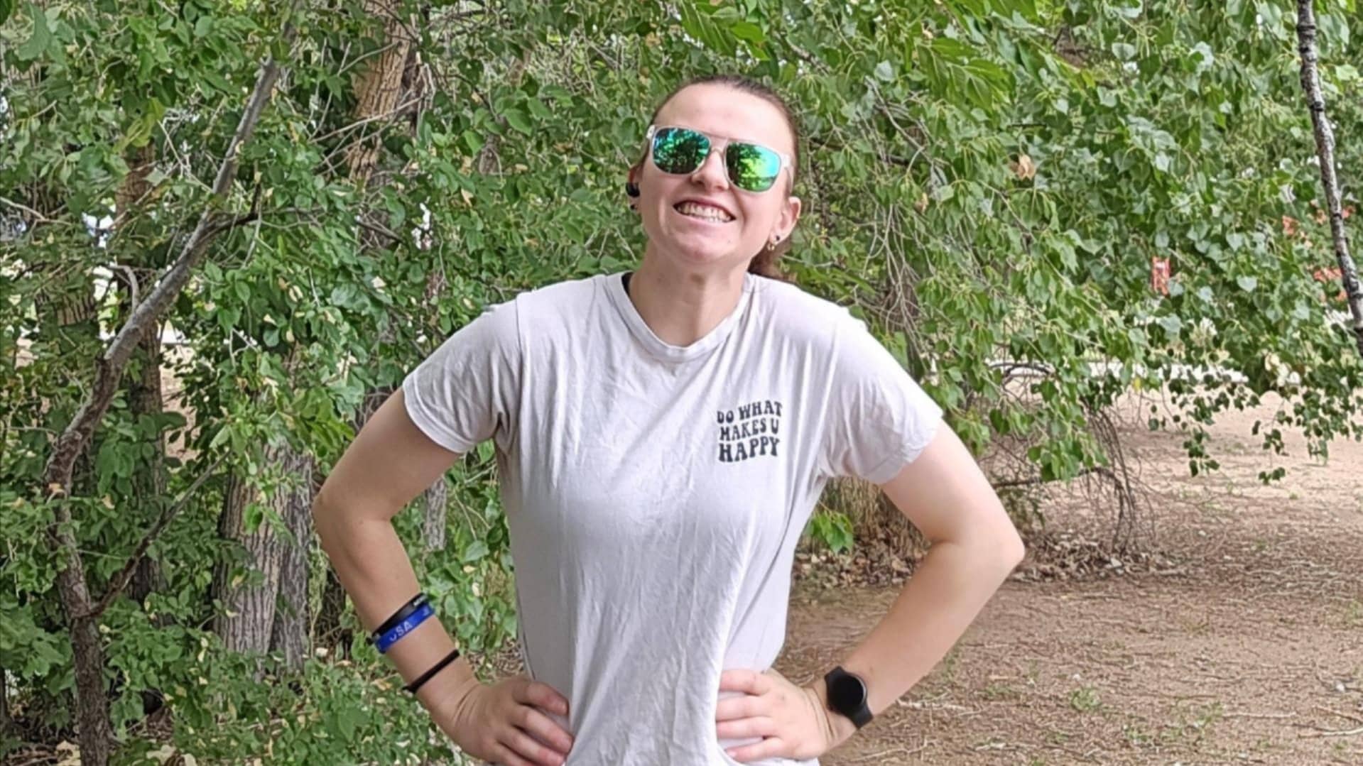 A person wearing sunglasses and a white t-shirt stands outdoors in front of green trees, smiling with hands on hips.