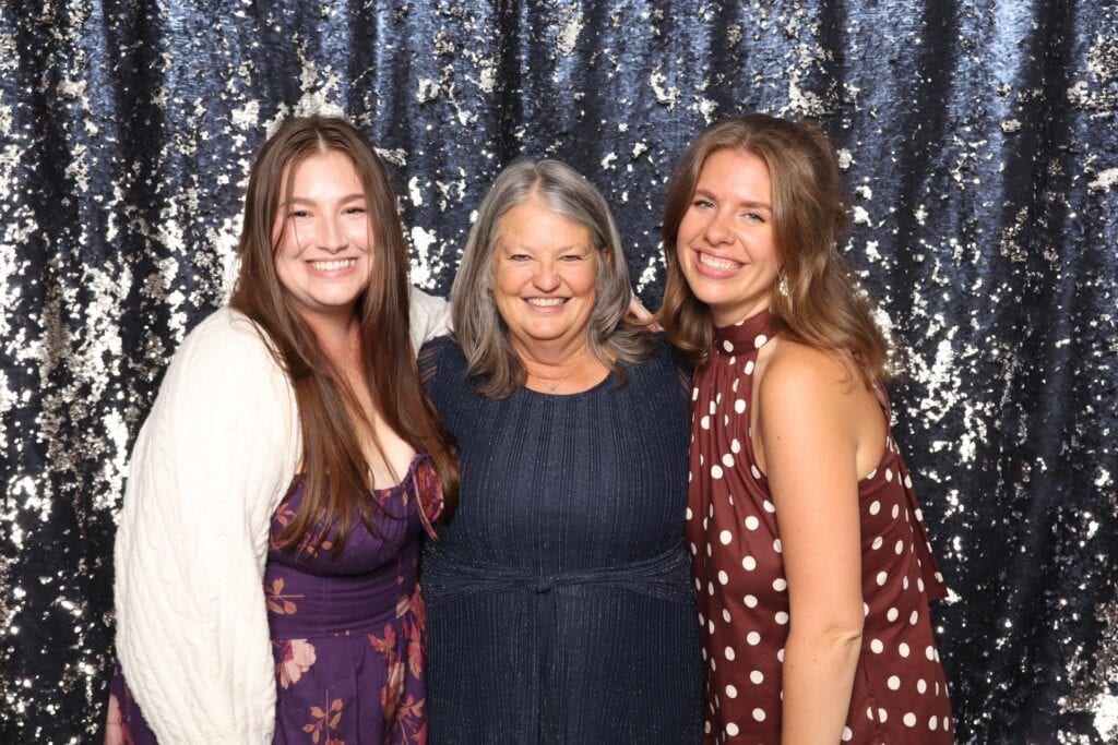 Three women smiling and posing together in front of a sparkly, dark blue sequin backdrop.