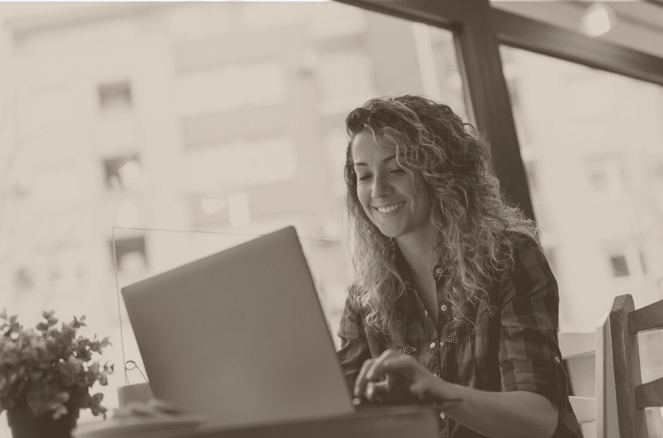 A woman sits at a table near a window, smiling while using a laptop. A small potted plant is on the table beside her.