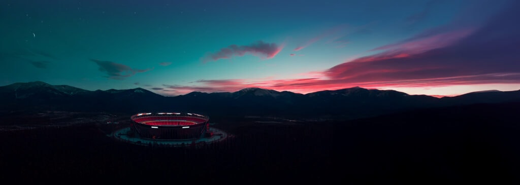 A large, illuminated stadium sits surrounded by forest with mountains and a colorful sunset sky in the background.