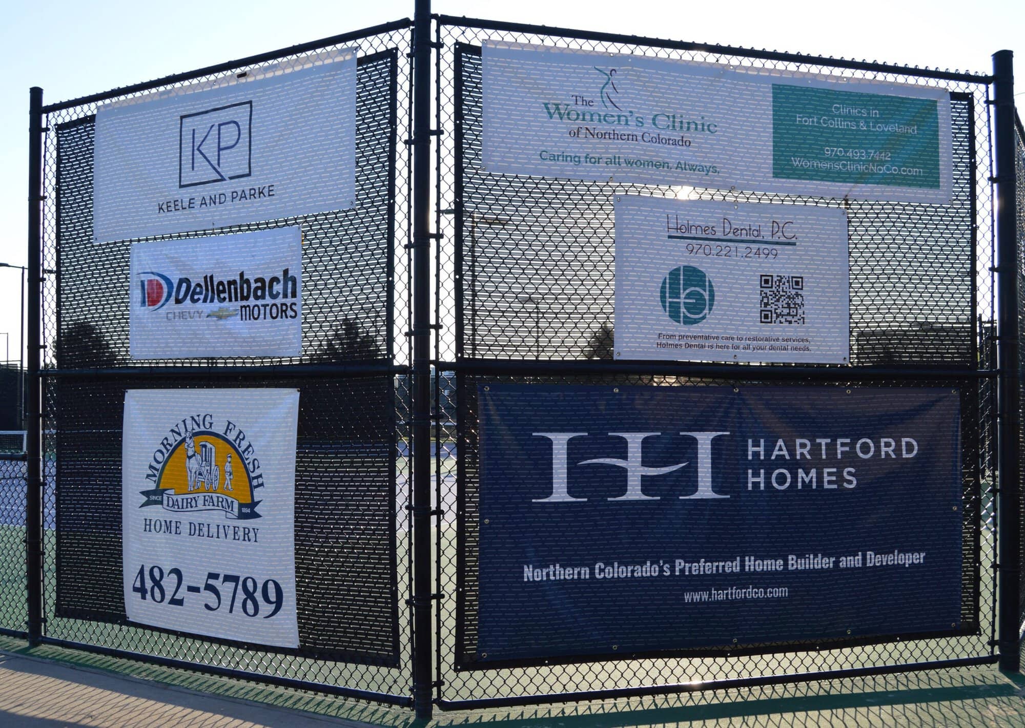 Banners displaying various business advertisements are attached to a black chain-link fence surrounding a tennis or sports court.