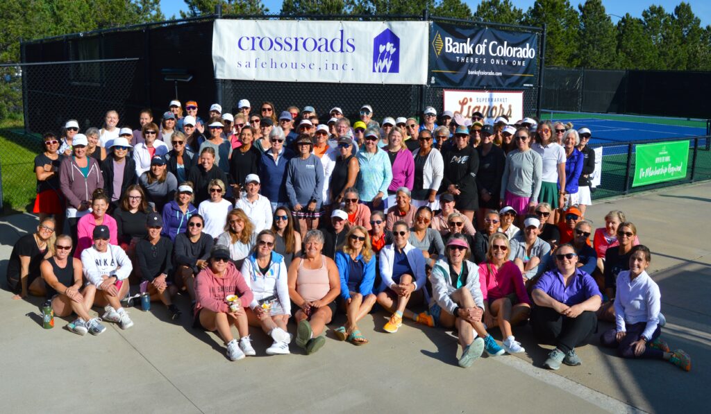 Large group of people posing together outdoors in front of tennis courts, wearing sportswear and sunglasses, with sponsor banners displayed behind them.