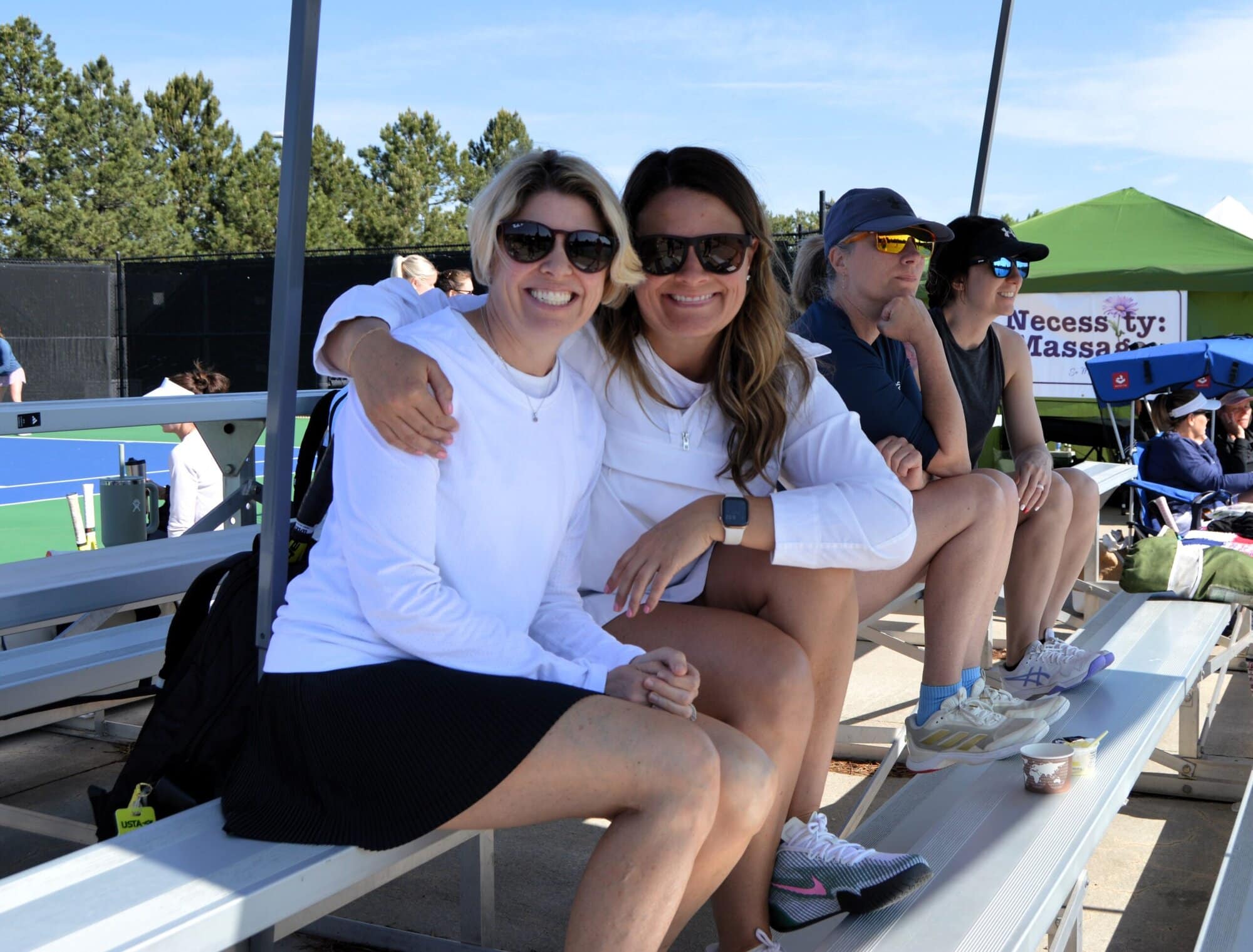 Two women sit together smiling on bleachers at an outdoor tennis event, with other people seated nearby and a tennis court in the background.