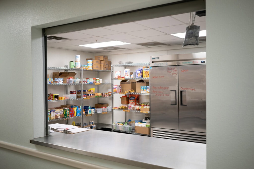 View through a service window into a stocked pantry with metal shelves, assorted food items, boxes, and a large stainless steel refrigerator with writing on the doors.