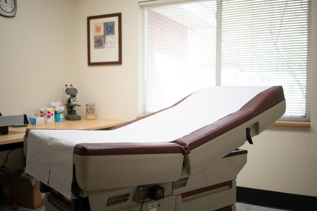 An examination table with a white paper cover in a medical office, next to a desk with medical supplies and a microscope, with sunlight coming through window blinds.