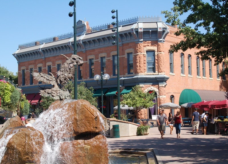 A brick building with green awnings stands behind a fountain featuring an eagle sculpture; people walk along the sunny sidewalk.
