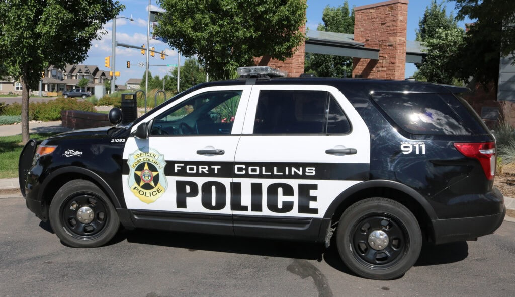 A Fort Collins Police SUV is parked on a street, with police insignia and "911" visible on the side. Trees and buildings are in the background.