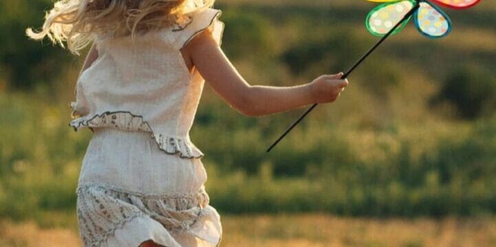 A young girl in a white dress runs through a field holding a colorful pinwheel, with the sun setting in the background.