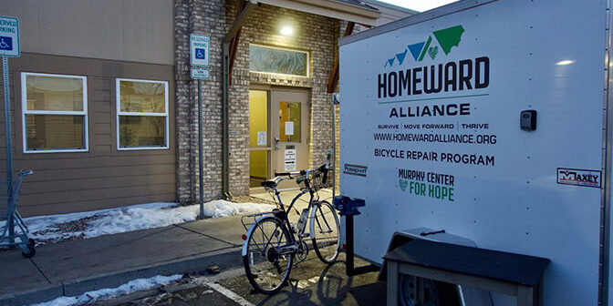 A bicycle repair trailer labeled "Homeward Alliance" is parked outside a building entrance with a bike stand and snow on the ground.