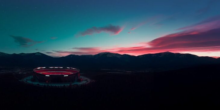 A large, illuminated stadium sits surrounded by forest with mountains and a colorful sunset sky in the background.
