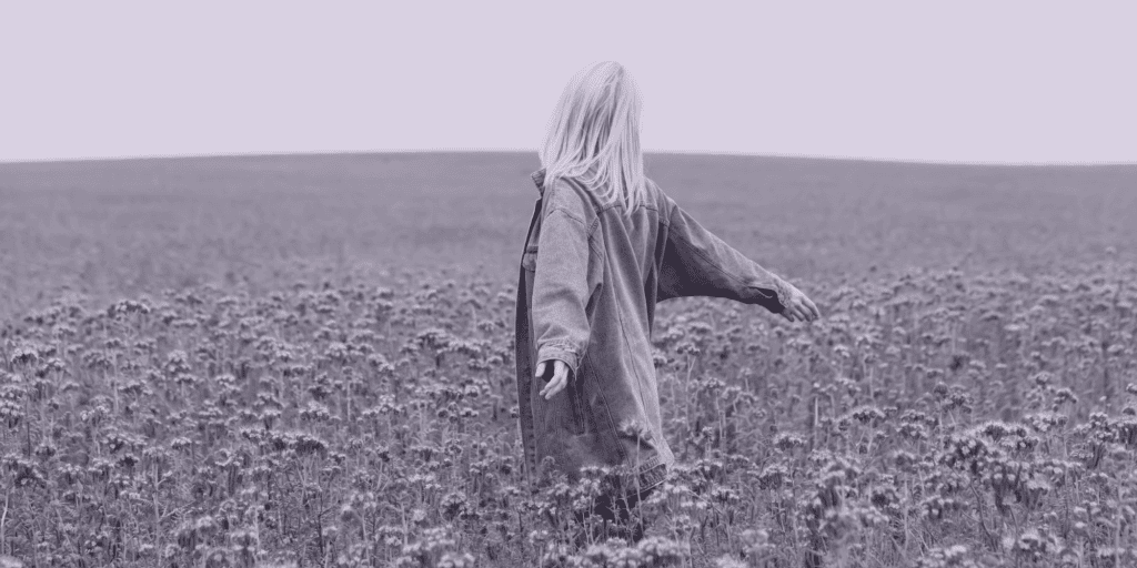 A person with long hair, wearing an oversized jacket, stands in a field of wildflowers with arms outstretched, facing away from the camera.