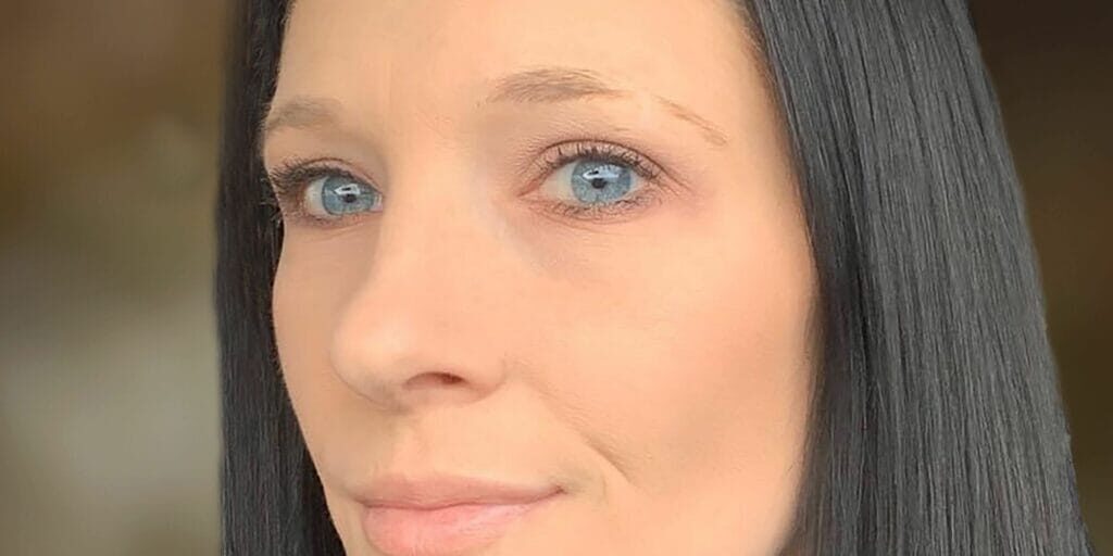 A woman with straight black hair and blue eyes looks at the camera with a neutral expression against a blurred background.