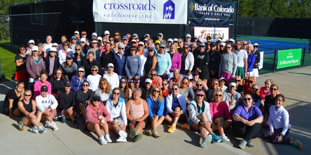 Large group of people posing together outdoors in front of tennis courts, wearing sportswear and sunglasses, with sponsor banners displayed behind them.