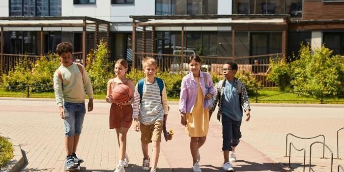 Five children walk together on a sidewalk in front of an apartment building; one rides a skateboard, another carries a basketball.