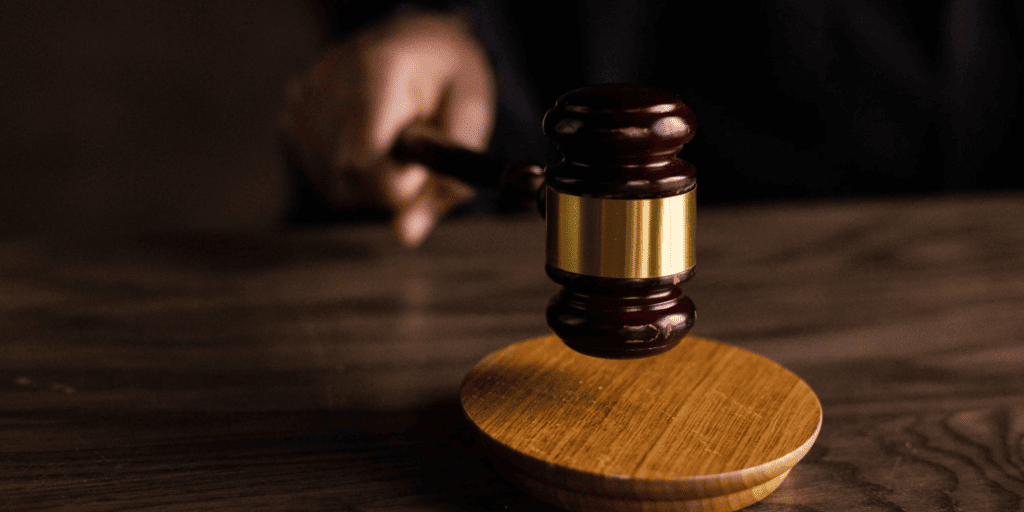 A judge’s hand holding a wooden gavel above a round block on a wooden table.