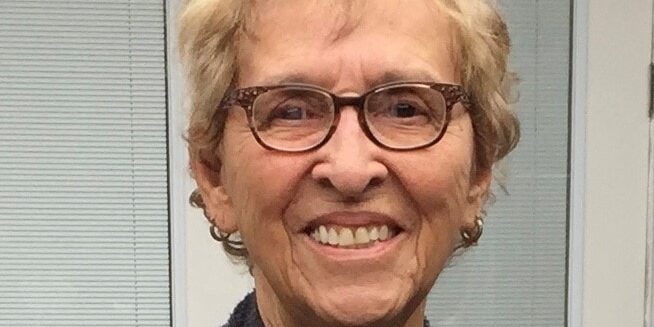 An older woman with short, light hair and glasses smiles at the camera, standing in front of a window with blinds.