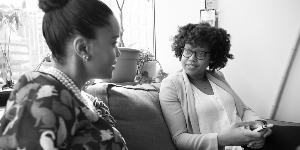 Two women sit on a couch having a conversation; one woman holds a phone and listens attentively while the other speaks.