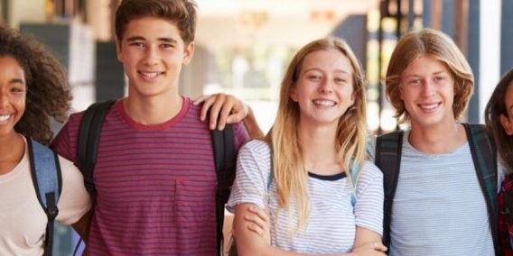 A group of seven empowering youth with backpacks stand closely together outdoors, smiling at the camera.