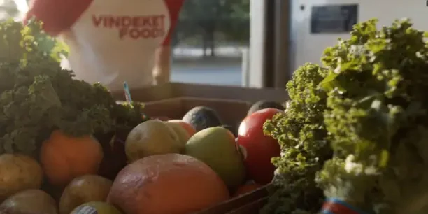 A box filled with assorted fresh fruits and vegetables sits on a table; a person in a "VINDEKIT FOOD" shirt stands in the background.